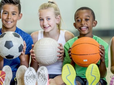 A group of children are indoors in their elementary school gym. They are sitting on the floor and getting ready to play sports. The kids are holding a basketball, a soccer ball, and three volleyballs.