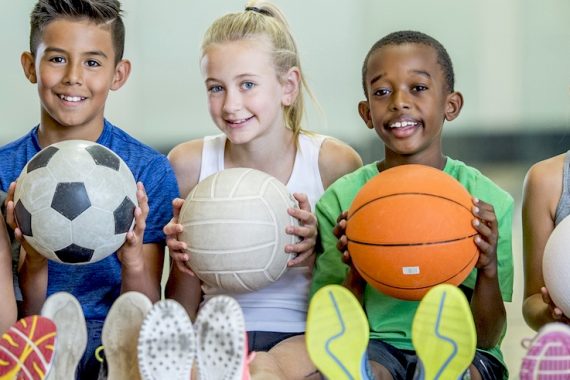 A group of children are indoors in their elementary school gym. They are sitting on the floor and getting ready to play sports. The kids are holding a basketball, a soccer ball, and three volleyballs.